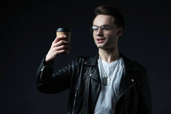 Excited stylish brutal man in biker jacket looking at coffee to go isolated on black — Stock Photo