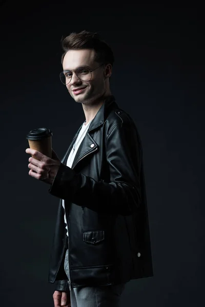 Seitenansicht des lächelnden, stylischen brutalen Mannes in Biker-Jacke mit Coffee to go auf Schwarz — Stockfoto