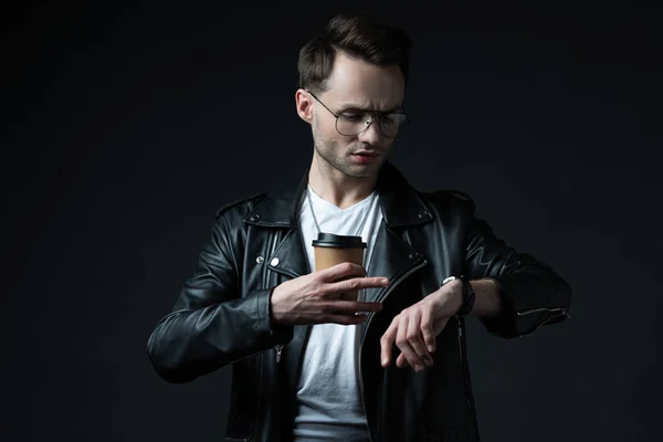 Elegante hombre brutal en chaqueta de ciclista mirando reloj de pulsera con café para ir aislado en negro - foto de stock