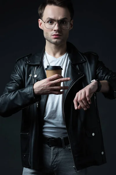 Stressed stylish brutal man in biker jacket with wristwatch and coffee to go isolated on black — Stock Photo
