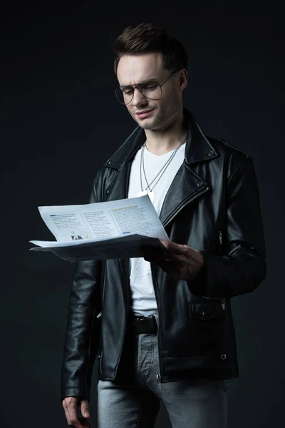 Smiling stylish brutal man in biker jacket with newspaper isolated on black — Stock Photo