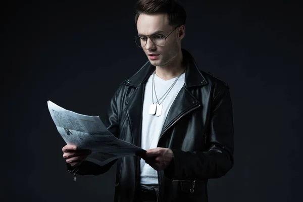 Shocked stylish brutal man in biker jacket with newspaper isolated on black — Stock Photo