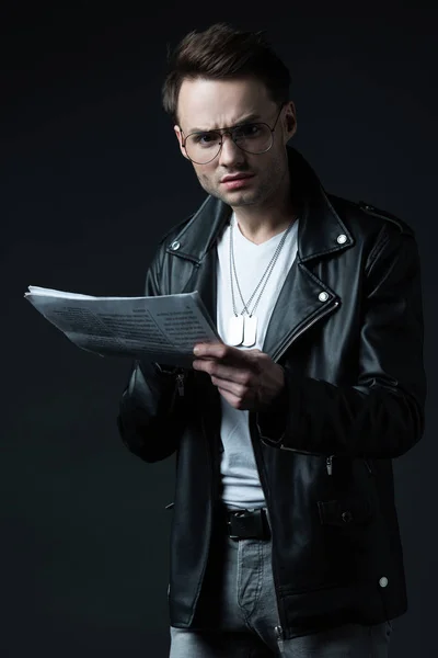 Confused stylish brutal man in biker jacket with newspaper isolated on black — Stock Photo