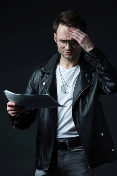 Reflexivo elegante hombre brutal en chaqueta de motociclista con periódico aislado en negro - foto de stock