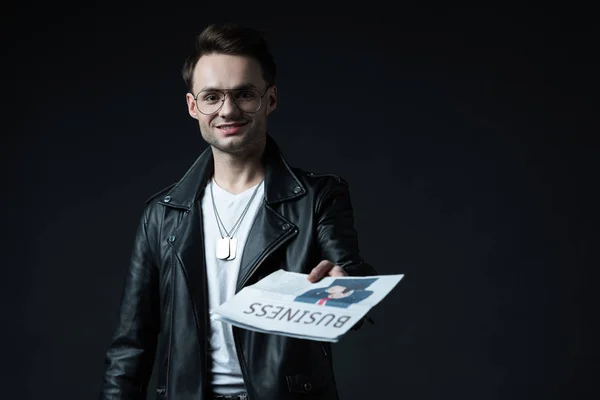 Selective focus of smiling stylish brutal man in biker jacket showing business newspaper isolated on black — Stock Photo