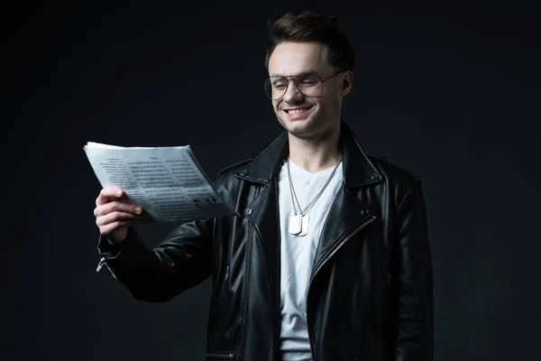 Smiling stylish brutal man in biker jacket with business newspaper isolated on black — Stock Photo