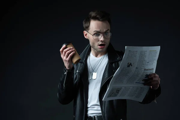 Shocked stylish brutal man in biker jacket with newspaper and coffee to go isolated on black — Stock Photo