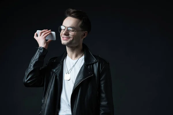 Sorrindo homem elegante em jaqueta de couro ouvindo mensagem de voz no smartphone isolado no preto — Fotografia de Stock