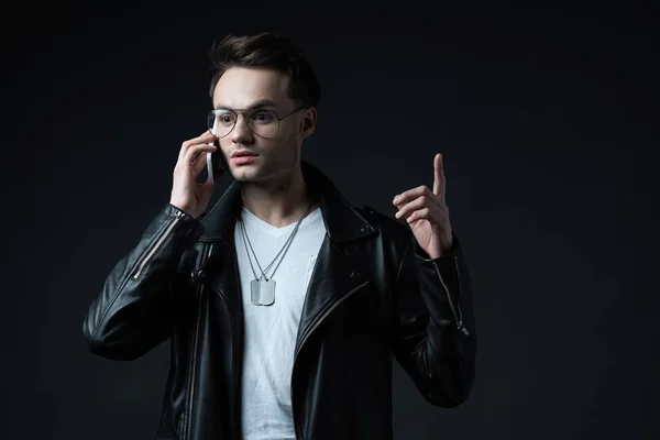 Hombre con estilo en chaqueta de cuero hablando en el teléfono inteligente y mostrando gesto idea aislado en negro - foto de stock