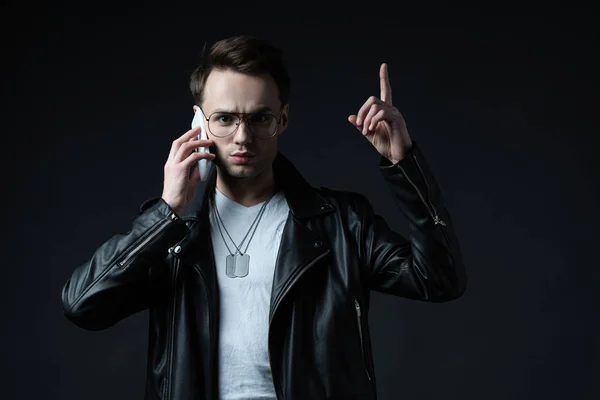 Tense stylish man in leather jacket talking on smartphone and showing idea gesture isolated on black — Stock Photo