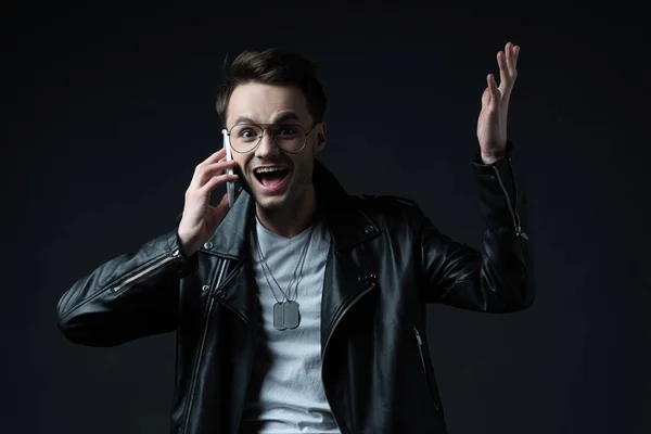 Feliz emocionado hombre con estilo en la chaqueta de cuero hablando en el teléfono inteligente aislado en negro - foto de stock