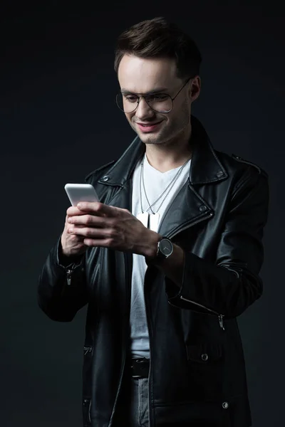 Улыбающийся стильный мужчина в кожаной куртке с помощью смартфона, изолированного на черном — стоковое фото