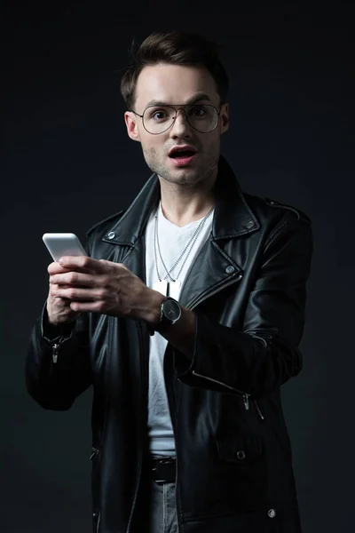 Impactado hombre con estilo en chaqueta de cuero usando teléfono inteligente aislado en negro - foto de stock