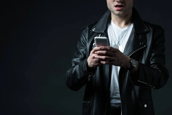 Vista recortada de impactado hombre elegante en chaqueta de cuero usando teléfono inteligente aislado en negro - foto de stock