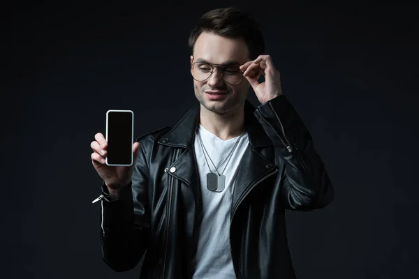 Улыбающийся стильный мужчина в кожаной куртке представляет смартфон с чистым экраном, изолированным на черном — стоковое фото