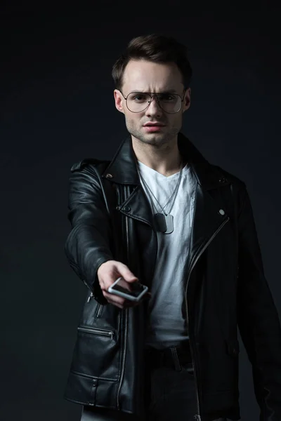 Hombre con estilo preocupado en chaqueta de cuero dando teléfono inteligente aislado en negro - foto de stock