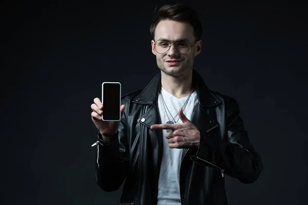Sonriente hombre elegante en chaqueta de cuero apuntando con el dedo al teléfono inteligente con pantalla en blanco aislado en negro - foto de stock