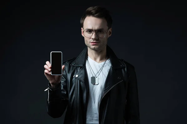 Hombre con estilo preocupado en chaqueta de cuero presentando teléfono inteligente con pantalla en blanco aislado en negro - foto de stock