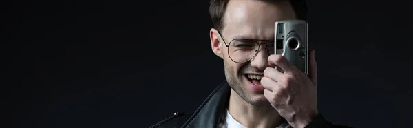 Happy stylish brutal man in biker jacket taking picture on film camera isolated on black, panoramic shot — Stock Photo