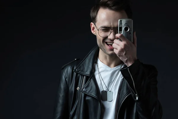 Feliz hombre brutal con estilo en la chaqueta del motorista tomando la foto en la cámara de cine aislado en negro - foto de stock