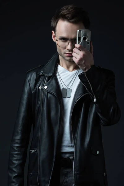 Serious stylish brutal man in biker jacket taking picture on film camera isolated on black — Stock Photo