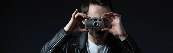 Stylish brutal man in biker jacket taking picture on film camera with open mouth isolated on black, panoramic shot — Stock Photo