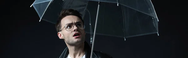 Shocked stylish brutal man in biker jacket with umbrella isolated on black, panoramic shot — Stock Photo