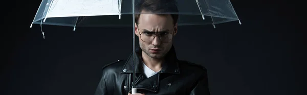 Angry stylish brutal man in biker jacket with umbrella isolated on black, panoramic shot — Stock Photo