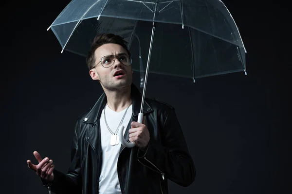 Shocked stylish brutal man in biker jacket looking at umbrella isolated on black — Stock Photo