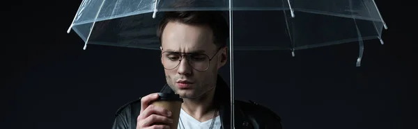 Worried stylish brutal man in biker jacket with umbrella and coffee to go isolated on black, panoramic shot — Stock Photo