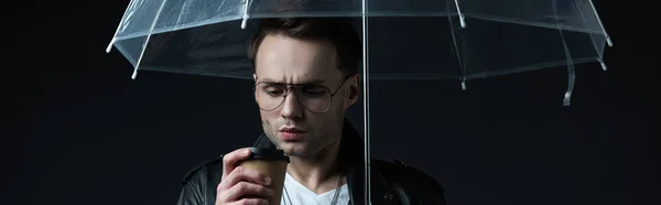 Preocupado hombre brutal con estilo en chaqueta de ciclista con paraguas y café para ir aislado en negro, tiro panorámico - foto de stock