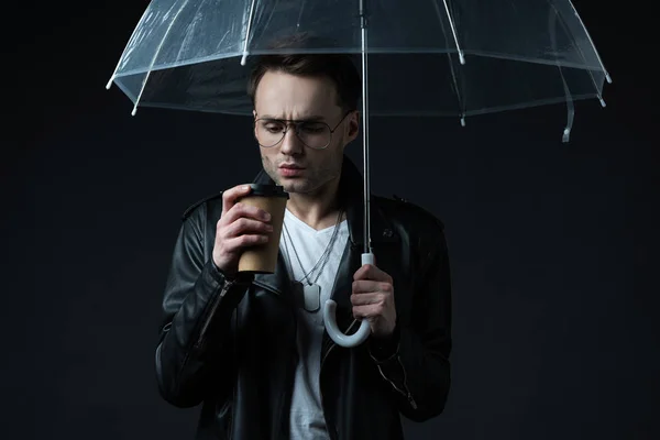 Worried stylish brutal man in biker jacket with umbrella and coffee to go isolated on black — Stock Photo
