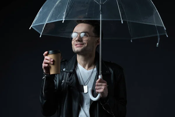 Sonriente elegante hombre brutal en chaqueta de motociclista con paraguas y café para ir aislado en negro - foto de stock