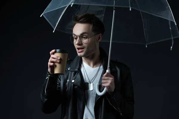 Shocked stylish brutal man in biker jacket with umbrella and coffee to go isolated on black — Stock Photo
