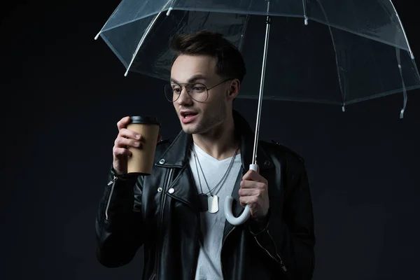 Schockierter brutaler Mann in Motorradjacke mit Regenschirm und Coffee to go auf schwarz — Stockfoto