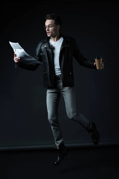 Shocked stylish brutal man in biker jacket levitating with paper cup and newspaper isolated on black — Stock Photo