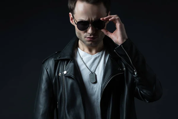 Stylish brutal man posing in biker jacket and sunglasses isolated on black — Stock Photo