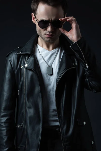 Stylish brutal man posing in biker jacket and sunglasses isolated on black — Stock Photo