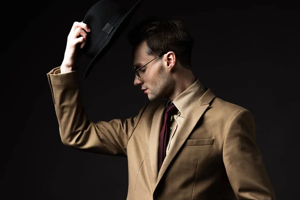 Elegant man in beige suit and eyeglasses holding hat isolated on black — Stock Photo