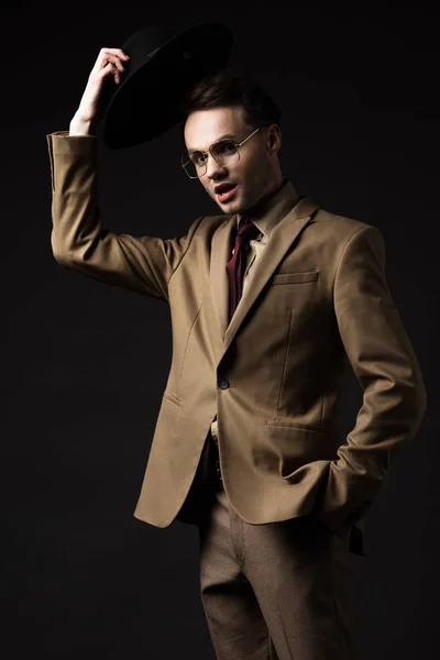 Impactado hombre elegante en traje beige y gafas con sombrero aislado en negro - foto de stock