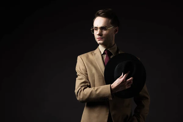 Hombre elegante pensativo en traje beige y gafas con sombrero aislado en negro - foto de stock