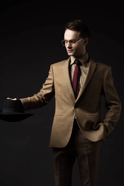 Sorrindo homem elegante em terno bege e óculos segurando chapéu isolado em preto — Fotografia de Stock