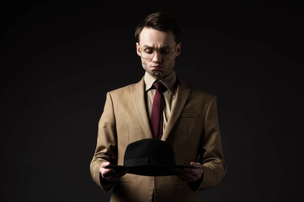 Elegant man in beige suit and eyeglasses holding hat and grimacing isolated on black — Stock Photo