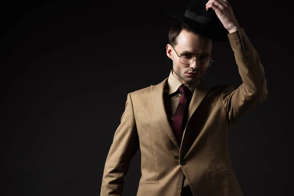 Hombre elegante en traje beige y anteojos que se ponen sombrero aislado en negro - foto de stock