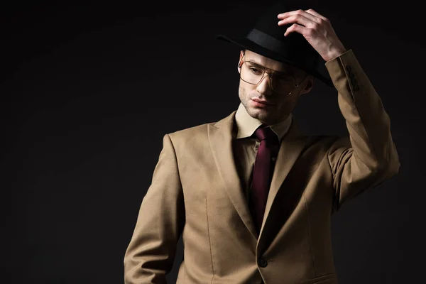 Hombre elegante en traje beige y anteojos que se ponen sombrero aislado en negro - foto de stock