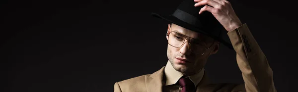 Hombre elegante en traje beige y anteojos que se ponen sombrero aislado en negro, tiro panorámico - foto de stock