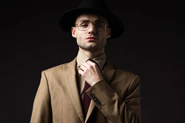Élégant homme en costume beige, chapeau et lunettes de fixation cravate isolé sur noir — Photo de stock