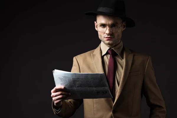 Hombre serio elegante en traje beige, sombrero y gafas con periódico aislado en negro - foto de stock