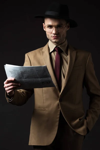 Hombre serio elegante en traje beige, sombrero y gafas con periódico aislado en negro - foto de stock