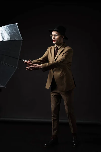 Shocked elegant man in beige suit, hat and eyeglasses holding umbrella on black — Stock Photo